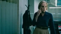 Mulher loira séria atende ao telefone num escritório escuro. Veste blusa azul e saia. Cabide com casaco ao fundo.