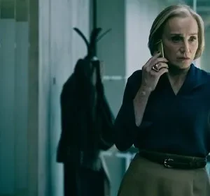 Mulher loira séria atende ao telefone num escritório escuro. Veste blusa azul e saia. Cabide com casaco ao fundo.