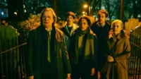 Um grupo de seis pessoas em um cemitério noturno. Todos parecem tensos sob a iluminação verde-escura do ambiente misterioso.