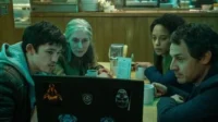 Um grupo diverso de quatro pessoas em um diner olham atentamente para um laptop. As expressões sérias criam um clima de foco e mistério.