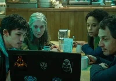 Um grupo diverso de quatro pessoas em um diner olham atentamente para um laptop. As expressões sérias criam um clima de foco e mistério.