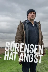 Um homem de gorro e jaqueta olha sério para o horizonte em campo verde. Céu nublado e ovelhas ao fundo criam clima sombrio.