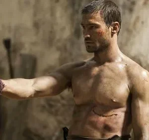 Homem musculoso e ferido, sem camisa, segura uma espada de madeira. Fundo rústico. Clima de luta e determinação.