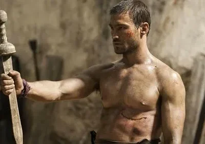 Homem musculoso e ferido, sem camisa, segura uma espada de madeira. Fundo rústico. Clima de luta e determinação.