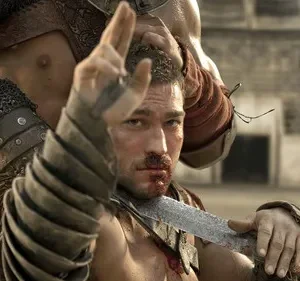 Gladiador sangrento, com espada, ergue a mão desafiadoramente. Guerreiro o segura pelo cabelo numa arena. Cena tensa e dramática.