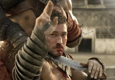 Gladiador sangrento, com espada, ergue a mão desafiadoramente. Guerreiro o segura pelo cabelo numa arena. Cena tensa e dramática.