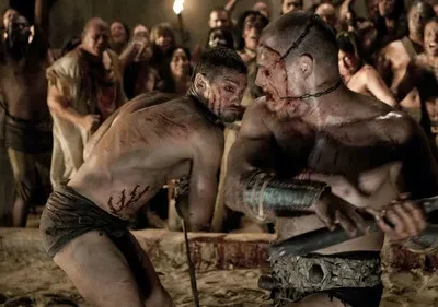 Dois gladiadores feridos e ensanguentados lutam na areia de uma arena. Multidão gritando na penumbra. Clima violento.