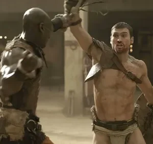 Dois gladiadores em combate feroz em arena poeirenta. Um guerreiro com escudo levanta arma, outro golpeia. Clima intenso e brutal.