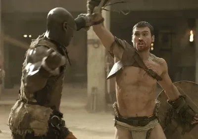 Dois gladiadores em combate feroz em arena poeirenta. Um guerreiro com escudo levanta arma, outro golpeia. Clima intenso e brutal.