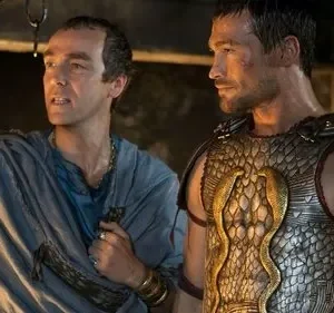 Dois homens, um de toga e outro em armadura escamada com cobras douradas, observam com seriedade. Cenário escuro e antigo.