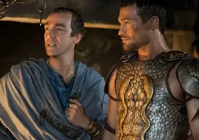 Dois homens, um de toga e outro em armadura escamada com cobras douradas, observam com seriedade. Cenário escuro e antigo.