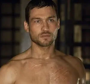 Homem musculoso, sem camisa, com olhar sério e cicatrizes no peito. Fundo escuro com grades. Clima de luta e resiliência.