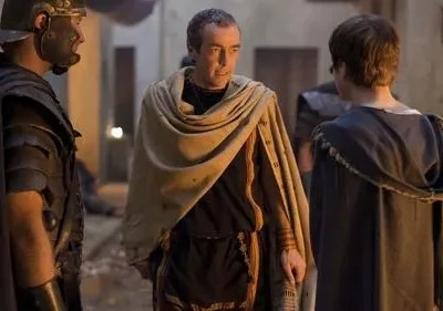 Três figuras em trajes antigos. Um soldado romano e um homem de toga conversam com uma pessoa de costas numa rua escura.