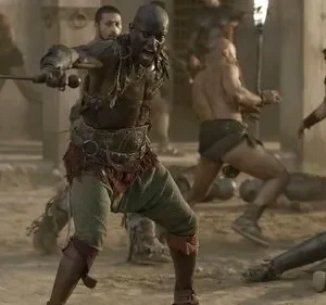 Um gladiador tribal raivoso empunha uma espada numa arena poeirenta, com soldados e corpos caídos ao redor. Clima tenso.