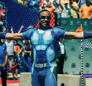 Homem negro em traje de super-herói azul/branco e óculos, braços abertos, com tala no braço esquerdo, celebrando em corrida.