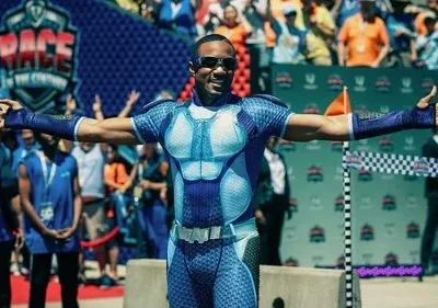 Homem negro em traje de super-herói azul/branco e óculos, braços abertos, com tala no braço esquerdo, celebrando em corrida.