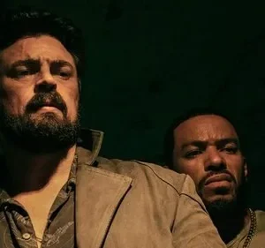 Homem barbudo com casaco marrom e cicatriz no rosto, olhando sério. Outro homem espreita por trás na penumbra verde.