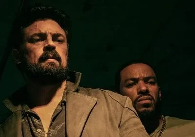 Homem barbudo com casaco marrom e cicatriz no rosto, olhando sério. Outro homem espreita por trás na penumbra verde.