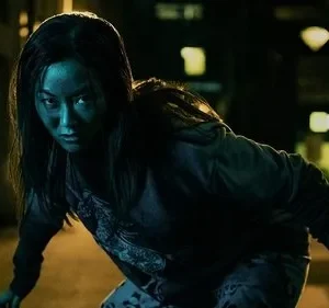 Mulher de rosto azul-esverdeado e olhar fixo, agachada em um beco escuro à noite. Uma silhueta ao fundo. Clima tenso.