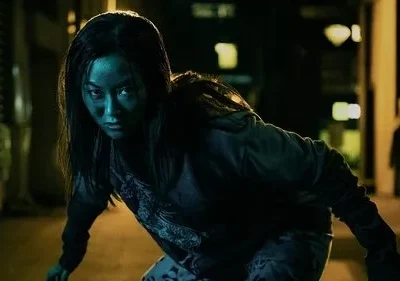 Mulher de rosto azul-esverdeado e olhar fixo, agachada em um beco escuro à noite. Uma silhueta ao fundo. Clima tenso.