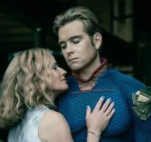 Mulher loira abraça homem de traje azul e dourado, que olha pensativo. Fundo escuro. Clima íntimo, mas melancólico.