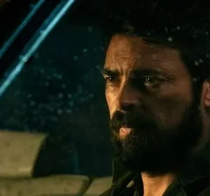 Homem barbudo e sério dentro de um carro, olhando através de um para-brisa molhado pela chuva à noite. Clima sombrio.