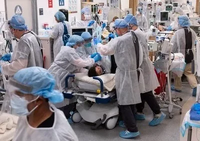 Muitos profissionais de saúde em EPIs brancos trabalham freneticamente cuidando de pacientes em uma UTI lotada com equipamentos médicos. Cena urgente.