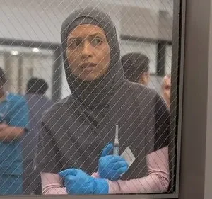 Mulher de hijab, luvas e scrubs segura seringa, com expressão séria, olhando por janela gradeada. Fundo desfocado.