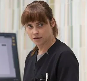 Mulher de uniforme médico preto com franja, olhar preocupado para o lado, em escritório médico com monitor e cortinas.