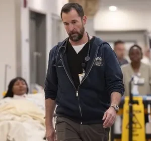 Médico barbudo com estetoscópio caminha sério em corredor de hospital. Paciente em maca e equipe médica ao fundo. Ambiente agitado.