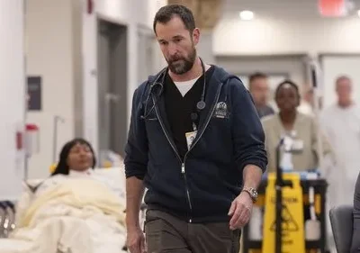 Médico barbudo com estetoscópio caminha sério em corredor de hospital. Paciente em maca e equipe médica ao fundo. Ambiente agitado.