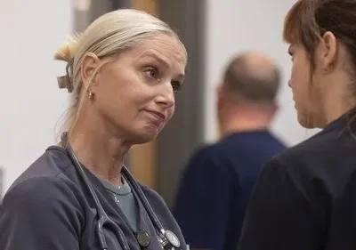 Uma enfermeira loira com estetoscópio e expressão séria conversa com outra pessoa em um ambiente médico.