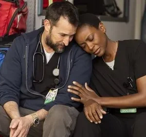 Dois médicos exaustos, um homem barbudo e uma mulher, se apoiam com tristeza no interior de uma ambulância.