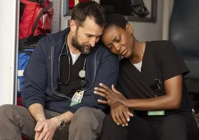 Dois médicos exaustos, um homem barbudo e uma mulher, se apoiam com tristeza no interior de uma ambulância.