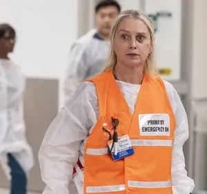 Mulher loira séria com colete de emergência em corredor de hospital. Outros profissionais de saúde desfocados ao fundo.