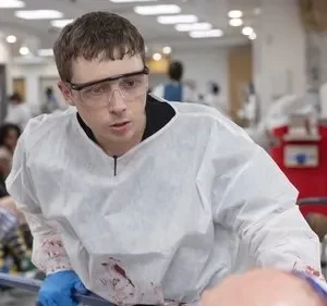 Médico jovem de óculos e roupa protetora ensanguentada em hospital caótico. Vários feridos no fundo, clima de urgência.