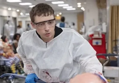 Médico jovem de óculos e roupa protetora ensanguentada em hospital caótico. Vários feridos no fundo, clima de urgência.