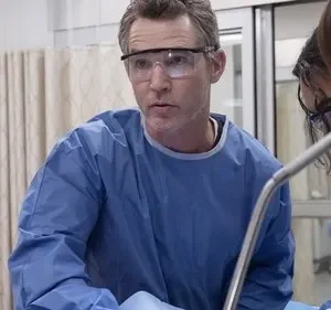 Três profissionais de saúde em azul e óculos, curvados e focados num procedimento médico. Cena tensa em um hospital.