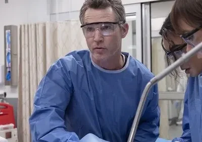 Três profissionais de saúde em azul e óculos, curvados e focados num procedimento médico. Cena tensa em um hospital.