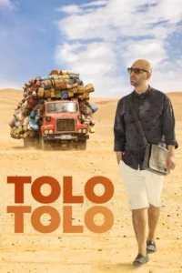 Homem de óculos escuros e shorts brancos caminha no deserto. Ao fundo,um caminhão vermelho sobrecarregado de malas.
