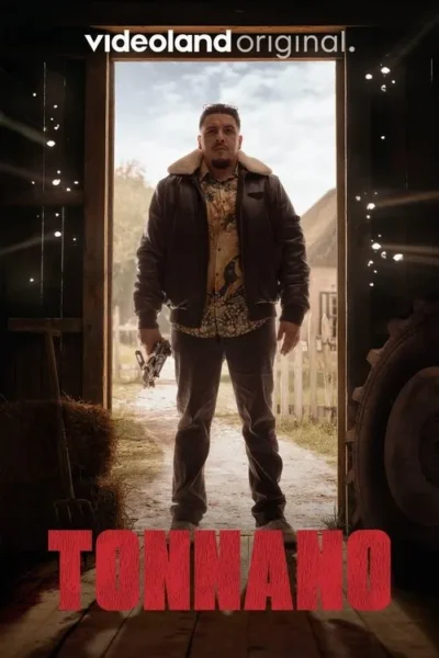 Homem sério de jaqueta e camisa estampada, com arma, na porta escura de um celeiro que se abre para um campo rural.