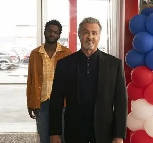 Homem mais velho de terno sorri ao lado de jovem sério de jaqueta. Fundo: concessionária e balões azul, vermelho e branco.