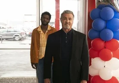 Homem mais velho de terno sorri ao lado de jovem sério de jaqueta. Fundo: concessionária e balões azul, vermelho e branco.