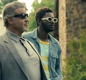 Dois homens de óculos de sol. Um grisalho de terno cinza e um jovem de jaqueta azul com óculos brancos, ao ar livre.