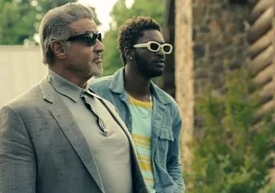 Dois homens de óculos de sol. Um grisalho de terno cinza e um jovem de jaqueta azul com óculos brancos, ao ar livre.