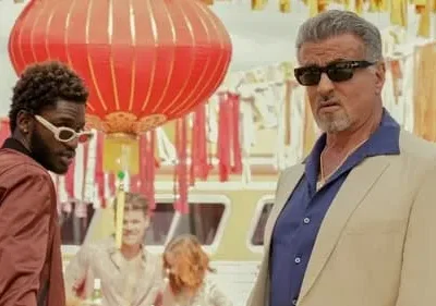 Homem sério de óculos e terno claro ao lado de jovem estiloso de óculos brancos. Fundo com lanterna vermelha e decorações festivas.