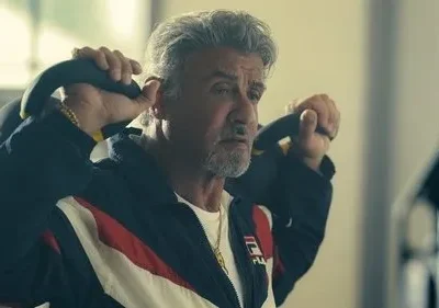 Homem de cabelo grisalho e barba, agasalho listrado, ergue dois kettlebells com expressão séria em uma academia.