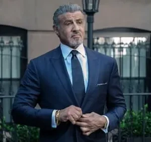 Homem de terno azul escuro e cabelo grisalho ajusta o paletó, com olhar sério. Cenário urbano com edifício e poste de luz. Tom formal.