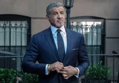 Homem de terno azul escuro e cabelo grisalho ajusta o paletó, com olhar sério. Cenário urbano com edifício e poste de luz. Tom formal.