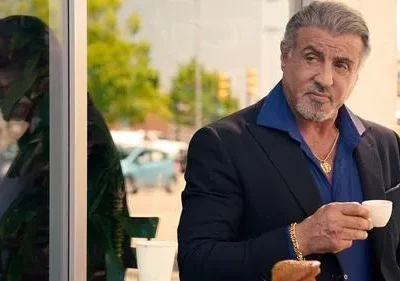 Homem grisalho de paletó escuro e camisa azul, colar dourado, segura uma xícara. Olhar sério, fundo urbano com reflexos.